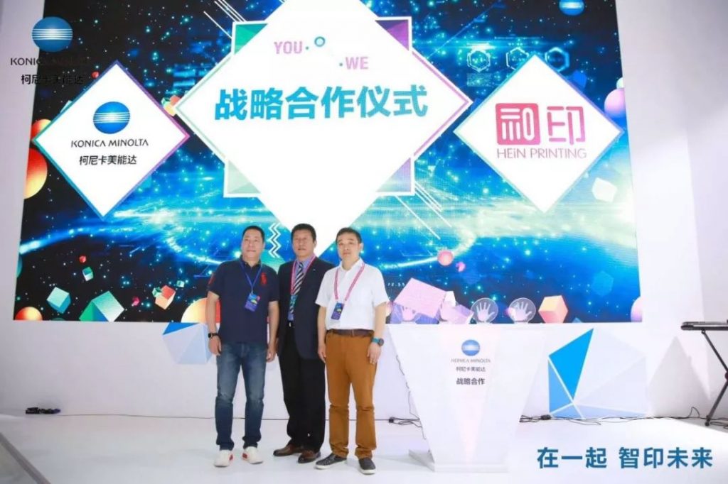 三名男子在柯尼卡美能达与和印标志布景前合影